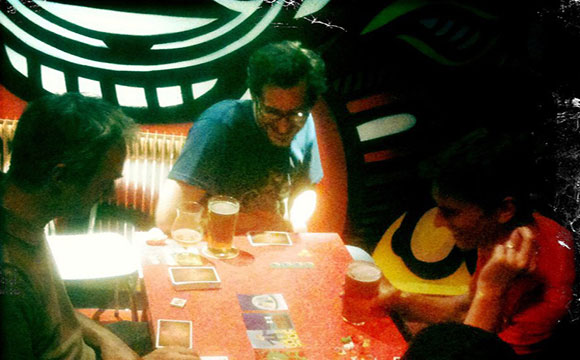 Trois personnes attablées à une partie de jeu de cartes, bières à la main